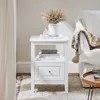 A white nightstand with books and a vase of dried flowers on top, next to a white sofa with a patterned throw blanket. Náhľad obrázku produktu