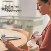 A woman is using a Philips AVENT hand breast pump while looking at her phone. The background shows a crib and a table with newspapers. Produktbild-Vorschau