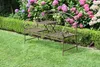 A garden bench with a small table in the middle, set in a garden with green grass and flowering bushes. Produktbild-Vorschau