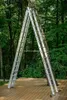 An aluminum ladder is set up in an A-frame configuration against a tree in a wooded area, on a wooden deck. Náhľad obrázku produktu