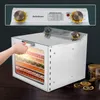 A hand is opening the door of a stainless steel dehydrator filled with dried fruits and vegetables. The dehydrator is on a wooden table, with some dried fruit slices scattered nearby. Podgląd zdjęcia produktu