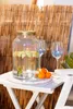 A beverage dispenser and wine glasses are set up on a small table outdoors, with a bamboo screen in the background. Produktbild-Vorschau