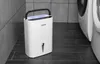 A white dehumidifier is placed on a dark tiled floor next to a washing machine, with gray tiled walls in the background. Náhľad obrázku produktu
