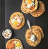 A top-down view of several small pancakes topped with various ingredients, arranged on a dark surface. Voir les images du produit
