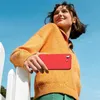 A young woman is holding a red smartphone case while leaning on a white fence outdoors. Produktbild-Vorschau