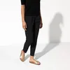 A person wearing black pants and a black top, standing and wearing gold Birkenstock sandals. Voir les images du produit
