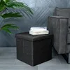 A dark gray fabric storage ottoman sits on a tiled floor next to a gray sofa. Two books are stacked on top of the ottoman. A large green plant is to the left of the ottoman. Náhled obrázku produktu