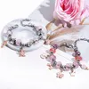 Two charm bracelets with pink beads and charms are displayed on a marble-like surface, with a large pink rose in the background. Produktbild-Vorschau
