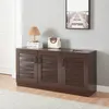 A dark brown shoe cabinet with three louvered doors is shown in a room with a light gray wall and a white door. A lamp, books, and a handbag are on top of the cabinet. Náhľad obrázku produktu