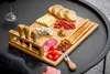 A charcuterie board with cheese, meats, bread, and nuts on a black table, with a glass of red wine in the background. Náhľad obrázku produktu