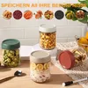 A collection of glass jars filled with various food items, displayed on a table with a textured cloth and a spoon. The background shows a tiled wall and a basket of fruit. Produktbild-Vorschau