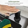 A hand is pressing down on a black soap dispenser next to a sink, with a sponge in the foreground. Produktbild-Vorschau