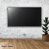 A black TV mounted on a white brick wall with a cable management system visible below. A potted plant is to the right of the TV. Náhled obrázku produktu