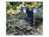 A black LED garden light with two lamps and a sensor is mounted among bushes and plants in a garden. Produktbild-Vorschau
