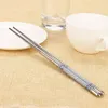 A pair of stainless steel chopsticks rests on a textured beige tablecloth, with a white plate, a clear glass, and a white cup in the background. Produktbild-Vorschau