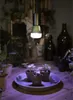 A grow light illuminates small seedlings in pots on a wooden surface, with other plants and bottles in the background. Anteprima dell’immagine prodotto