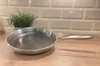 A stainless steel frying pan with a textured interior sits on a wooden countertop, with a potted plant and a tiled backsplash in the background. Produktbild-Vorschau
