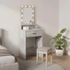A vanity table with a lighted mirror, a drawer, and a stool, set up in a bedroom next to a bed and a potted plant. Podgląd zdjęcia produktu