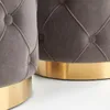 Close-up view of two upholstered ottomans with tufted detailing and gold bases. Produktbild-Vorschau