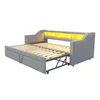 A grey upholstered daybed with a pull-out trundle bed extended, showing the wooden slats. The headboard has integrated yellow LED lighting. Produktbild-Vorschau