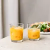 Two glasses of orange juice with ice on a counter, with a salad in the background. Produktbild-Vorschau