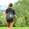 A young person walks away from the camera in a grassy, outdoor setting, wearing a black t-shirt and shorts, with a black drawstring backpack on. Produktbild-Vorschau