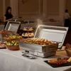 A buffet setup with various dishes, including a chafing dish with food, salads, and desserts. A person is visible in the background. Náhled obrázku produktu
