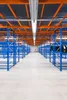 An empty warehouse aisle with rows of blue shelving units and orange overhead structures. Produktbild-Vorschau