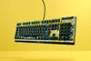 A gaming keyboard with a colorful, patterned design is shown floating against a bright yellow background. Produktbild-Vorschau