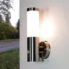 A modern stainless steel outdoor wall light with a motion sensor is mounted on a textured wall. The light is illuminated, casting a warm glow. To the left, a road and trees with autumn foliage are visible. Produktbild-Vorschau