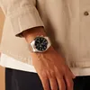 A close-up shot of a man's wrist wearing a silver chronograph watch with a black dial, as he is wearing a tan jacket. Podgląd zdjęcia produktu
