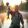 A man stands in a misty lake at sunrise, holding up a freshly caught fish. He is wearing fishing waders and a plaid shirt. Produktbild-Vorschau
