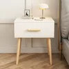 A white bedside table with a wooden drawer handle and legs, placed next to a bed. Náhľad obrázku produktu