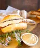 A sausage in a bun with sauce, served with herbs and a small bowl of sauce on a wooden board. Náhľad obrázku produktu