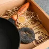 A cast iron pan and accessories are arranged in a cardboard box, cushioned with straw. Produktbild-Vorschau