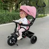 A baby sits in a pink tricycle outdoors. Produktbild-Vorschau