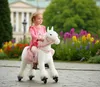 A young girl rides a white toy horse with pink accents in a park with flowers and a building in the background. Anteprima dell’immagine prodotto