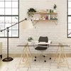 A modern home office setup with a glass-top desk supported by wooden trestle legs, a desk chair, a floor lamp, and shelves on a white brick wall. Produktbild-Vorschau