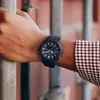 A person's arm wearing a black G-Shock watch, with a blurred background of industrial containers. Náhled obrázku produktu