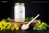 A jar of honey is displayed with yellow flowers and a wooden spoon. Náhled obrázku produktu