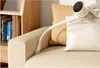A white handheld vacuum cleaner with a bent attachment cleaning a beige sofa with a yellow pillow behind it. A wooden bookshelf filled with books is visible on the left. Podgląd zdjęcia produktu