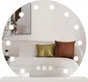 A round mirror with lights is placed on a shelf, reflecting a sofa with pillows and a framed picture. Náhled obrázku produktu