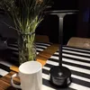 A black LED table lamp is placed on a table with a black and white striped tablecloth, next to a white mug and a vase of dried flowers. Produktbild-Vorschau