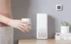 A person is holding a white cup next to a white Xiaomi air purifier on a wooden surface. A small digital thermometer is mounted on the wall behind the air purifier. Podgląd zdjęcia produktu