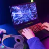 A person's hands are typing on a gaming laptop with RGB keyboard lighting, with headphones and a game controller visible on the desk. Náhľad obrázku produktu