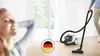 A woman relaxing while a person vacuums in the background, with a German engineering logo overlay. Voir les images du produit