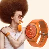 A person with a large afro hairstyle is holding an orange smartwatch in their mouth, with another orange smartwatch on their wrist. Voir les images du produit