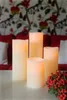 Four LED candles of varying heights are arranged on a white surface in front of a red sofa with a floral patterned wallpaper. Podgląd zdjęcia produktu