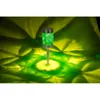 A green solar garden light with a patterned cylinder is illuminated, casting a green and yellow light pattern on the ground. Produktbild-Vorschau