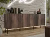 A modern wooden sideboard with ribbed doors is displayed in a living room setting, with decorative items on top and a neon light fixture on the wall. Produktbild-Vorschau